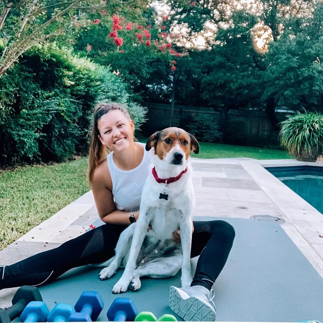 Woman in sneakers sitting with her dog on a large grey mat by a backyard pool, dumbbells in front