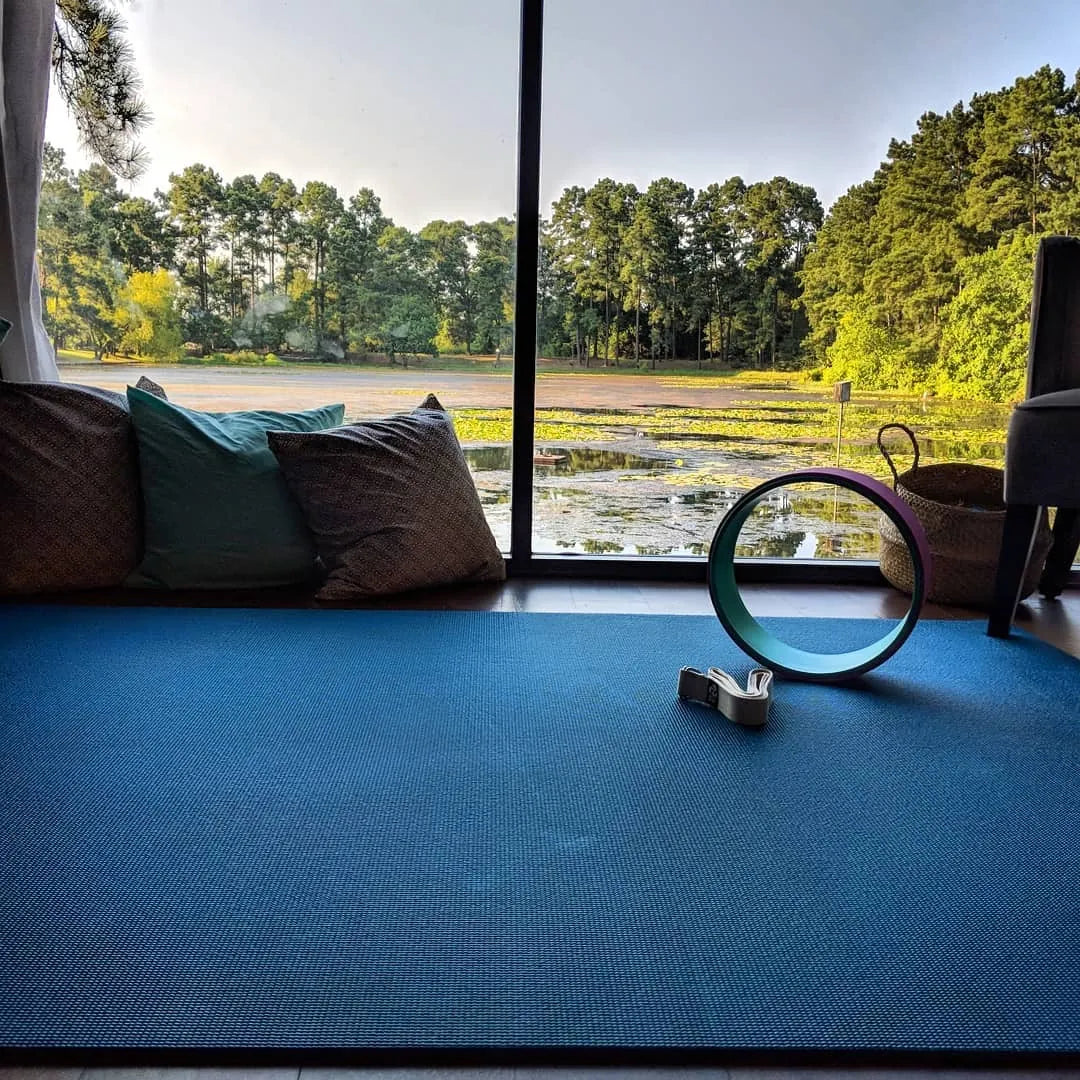 Teal yoga mat by a floor-to-ceiling window looking out on trees, with a yoga wheel
