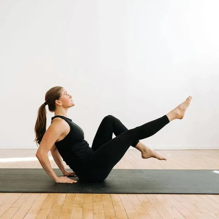 Barefoot woman doing a core exercise on a large black yoga mat in a bright studio with a wood floor